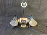 Front view of Ryobi bench grinder showing dual grinding wheels, safety shields, and flexible work light.