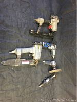 Five industrial pneumatic power tools arranged for overview: two nailers, impact wrench, grinder, and cutoff tool on dark backdrop.