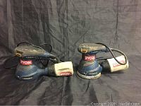 Two Ryobi electric sanders side by side on a black fabric backdrop, showing the main body and dust bags.