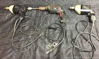 Three handheld corded power drills on black surface: two vintage style angled-handle drills and one modern black/red Skil drill, cords visible.