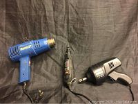 Three power tools laid out on black fabric: the Bostik 1200 heat gun on the left, Dremel Moto-Tool center, Black & Decker 12V impact wrench right.