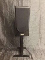 Front view of black Kenwood speaker on metal stand showing fabric cover and brand logo.
