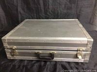 Closed view of grey rectangular flight case with metal framing, latches, and handle on black fabric background.