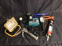 Photo showing all six tools on black background including yellow work light, black flashlight, cordless drill charger, grease gun, caulking gun, locking pliers