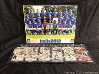 Photo showing Italia 2012 soccer team poster above and NASCAR 50 Years wooden plaque below, both propped against black background.