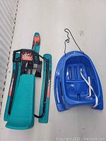 GT Snow Racer sled on the left in turquoise and black with red graphics, and blue plastic toboggan on the right with pull string and white strap.