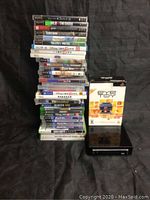 Tall stack of video game cases in various formats including Wii, PS3, PSP, Xbox 360, next to boxed PlayStation 2 Eye Toy camera and black Nintendo Wii console.