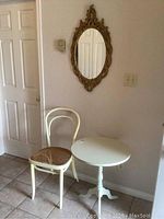 Full view of the small round table, white chair with rattan seat, and the oval mirror on the wall above them.