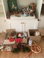 Wide view showing assorted Christmas decor spread out on a shelf and inside cardboard boxes on the floor including wreaths, figurines, gift boxes, and ribbons.