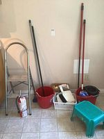 Photo showing two-step ladder, turquoise step stool, red bucket, fire extinguisher, sponge mop and broom inside a tiled room corner.