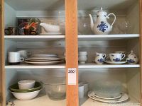 Full view of cabinet shelves with assorted kitchen dishes including teapot, clear serving glasses, ramekins, bowls, plates and cups.