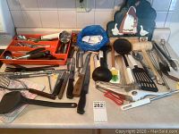 Photo of assorted cutlery and cooking utensils laid out on kitchen counter including forks, knives, spoons, ladles, spatulas, tongs, and more with an orange plastic flatware tray.