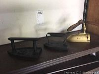Three antique flat irons displayed on a shelf, including two cast iron units and one brass and wood iron.