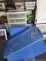 White five-drawer storage unit with transparent drawers and wheels in background. Large blue Rubbermaid storage bin with lid in foreground.