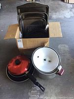 Box containing several metal roasting pans of different sizes and a black skillet with red lid and handle