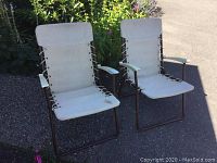 Photo of two vintage folding canvas chairs outdoors, showing white fabric seats and backs with laced sides, brown metal frames, and white plastic armrests.