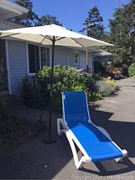 Outdoor lounge chair with white plastic frame and blue mesh fabric, shown in sunlight on pavement with greenery in background.