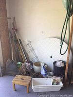 Overview showing various gardening tools and supplies including rakes, pitch fork, spade, metal tomato cage, pots, and bags of gardening amendments, with a small wooden bench.