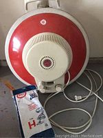 Front view of the round red and white canister vacuum with power cord and pack of original Hoover H vacuum bags on floor next to it.