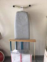 Vintage ironing board with blue metal legs, gray padded cover, and wooden rack for clothes or accessories.