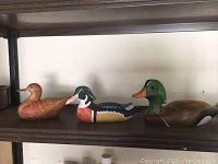 Three carved wooden ducks displayed on wooden shelf, showing various sizes and colors