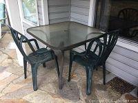Patio table with glass top and two green plastic chairs arranged on outdoor stone floor near a house exterior wall.