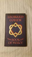 Front cover of the book showing purple cloth binding with gold foil title 'Leonard Cohen Book of Mercy' and emblem