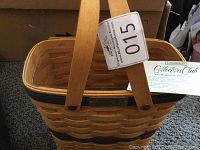 Front view showing woven wood basket with navy painted accents and two handles. Plastic liner visible inside. Metal badge for Longaberger Collectors Club fixed on upper side.