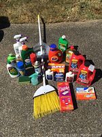 Photo of full cleaning lot assortment including broom, dustpan, various bottles of cleaning liquids, aerosol sprays, and sponges on outdoor pavement.