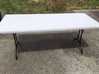 Full view of the folding table showing white top and black legs on concrete and grass background.