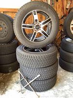 Set of four tires stacked, one mounted on alloy wheel on top clearly visible with 9 lug bolt holes and five-spoke design.