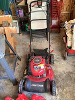 Front view of red Craftsman lawnmower with black grass catcher, placed in garage among other tools and items.