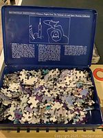 Opened metal box containing scattered space-themed jigsaw puzzle pieces with information about famous flights from the National Air and Space Museum printed on the lid.