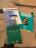 Photo showing three music books: Yamaha Performance Folio, Essential Elements, and Guitar 101 manual, placed on cardboard boxes.