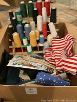 Box containing large wooden spools of sewing thread in multiple colors along with assorted fabrics and sewing pattern booklets.