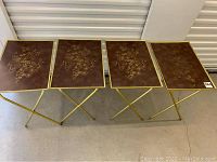 Four vintage TV trays with folded gold metal legs and brown tops with gold floral design, arranged in a row.