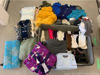 Assortment of sweaters, gloves, scarves, socks, and pajamas in and around an open suitcase, showcasing variety and colors of clothing and accessories