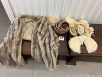 Wide shot of two light brown fur stoles, three belts, and a pair of cream moccasins placed on a wooden bench.