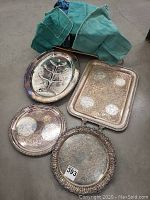Four silver-plated serving trays arranged on floor with two green fabric cutlery bags behind them