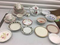 Array of assorted china pieces including cups, saucers, plates, and a teapot laid out on a table showing varied patterns and styles.