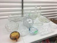 Wide view of assorted crystal and glass items including bowls, platters, ashtray, and decanters on white surface.