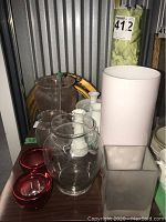 Group photo on shelf showing all vases and candle holders arranged close together, includes clear glass, milk glass, red votives, and pink vase.