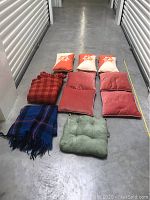 Assorted bedding items and throw pillows arranged on floor including plaid blankets and velvet cushion