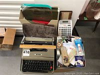 View of vintage Olivetti typewriter with cover, wooden ruler, large office calculator, and metal tray with office supplies including tape and pens.