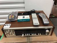 Front angled view of the Kemtron stereo receiver with control knobs, two remote controls, RCA AV cable, boxed pocket radio, and power cable on top.