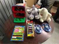 Full lot overview showing Matchbox cars, plush toys, coloring puzzle with markers, and handheld games on a table with LEGO container behind.