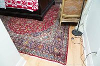 Rug under bed showing red field and surrounding navy border with floral motifs, edge wear visible