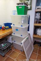 Stacked clear Rubbermaid storage bins with blue wheels and a foldable Safety 1st baby gate leaning against the stack, situated in a corner with wood shelving and some cans nearby.