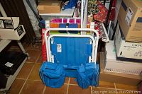 Folded blue chair with umbrella and two storage pouches on the arms, stacked with other outdoor chair and items around