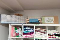 Wide view of shelf showing cutting mat, loom with blue weaving, original box, stacked fabric remnants and cushions